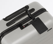 The Bigger Carry-On suitcase in Cloud Gray shown shown from above highlighting TSA-approved combination lock and recycled leather luggage tag