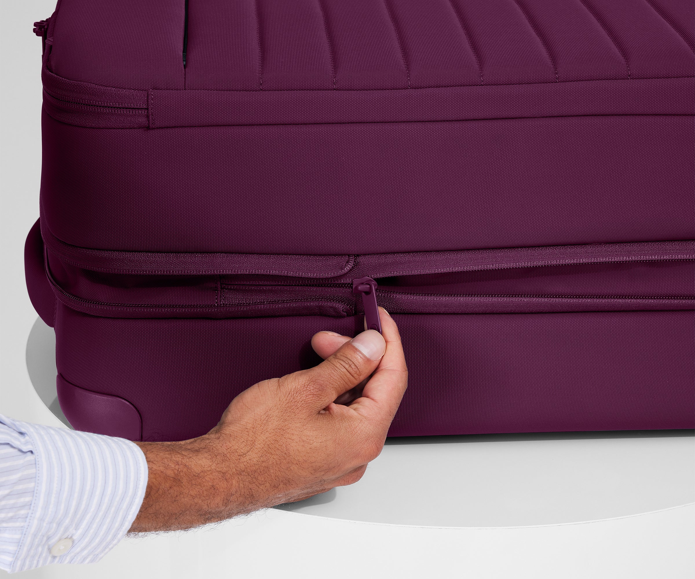 A person is unzipping The Softside Bigger Carry-On suitcase in Plum Purple using the wraparound zipper to access 1.75" (4.5 cm) of extra packing space